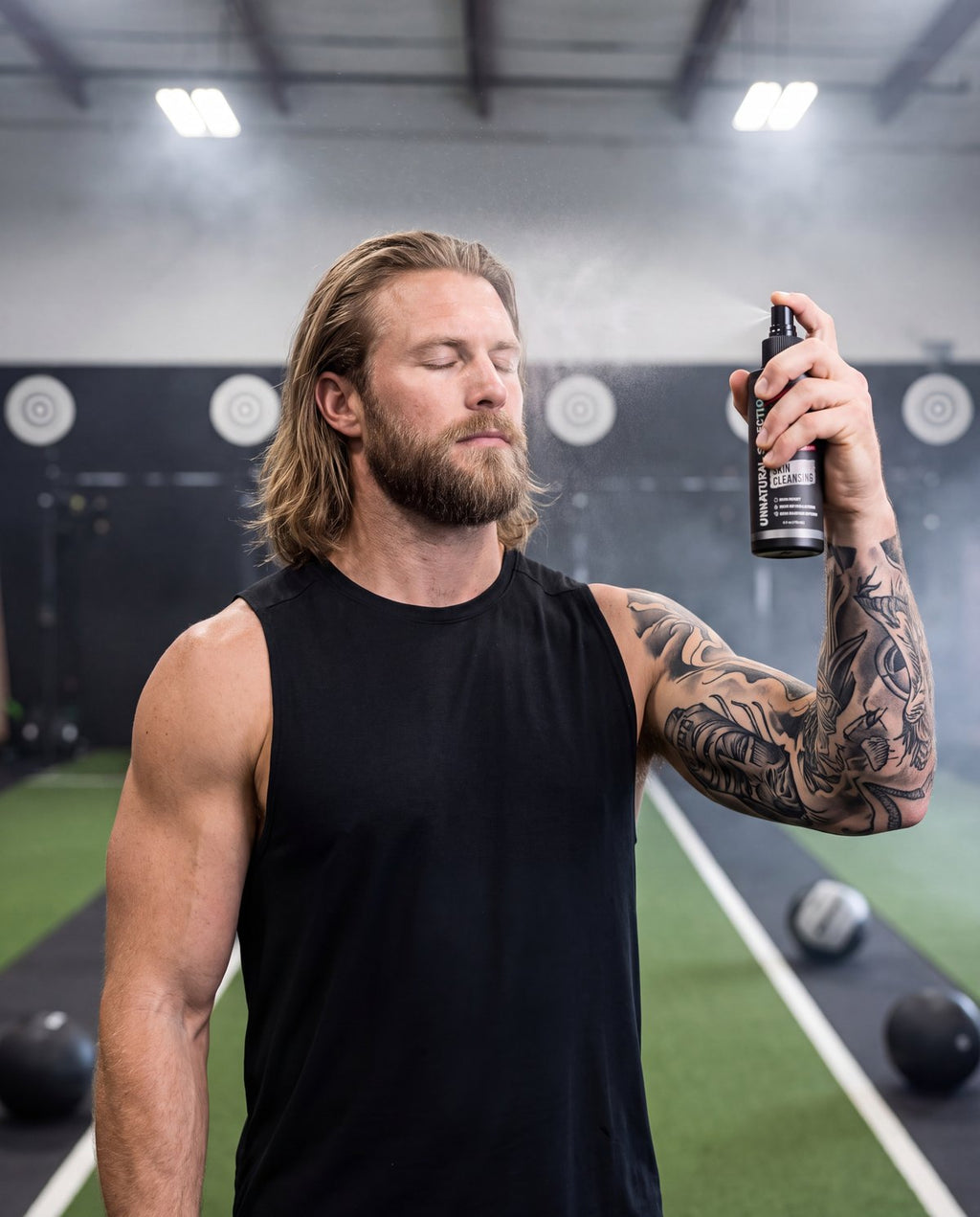 Tattooed athlete in a race arena spraying UNS skin cleansing spray on his face beside the wall-ball targets